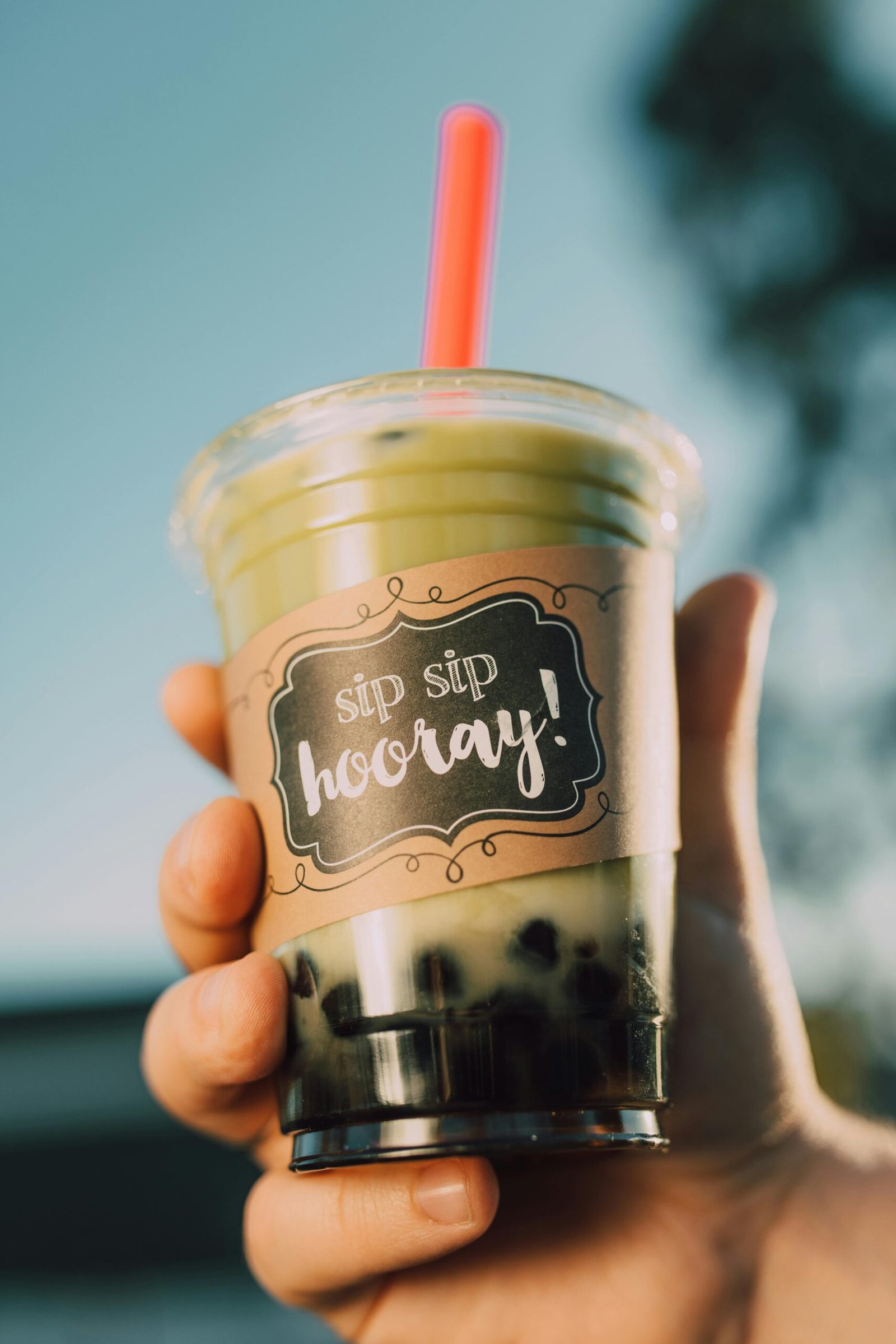 Close-up of a hand holding a matcha flavored boba tea outdoors with label saying 'sip sip hooray!'